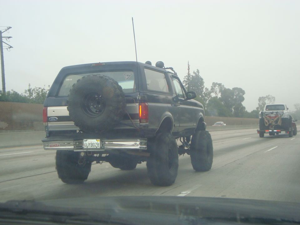 Lifted black Ford Bronco on highway, spare tire mounted on rear door. Another truck is visible ahead