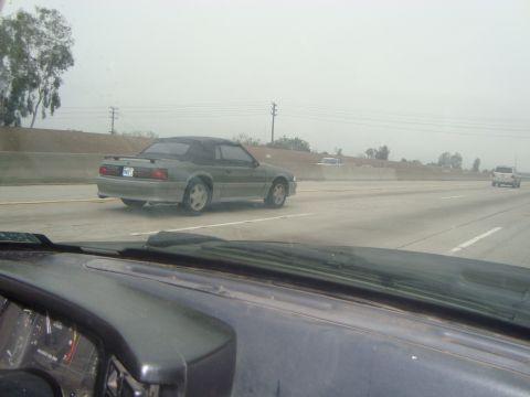 1986 Ford Bronco - A gray convertible Mustang is seen driving on a highway on a hazy day.