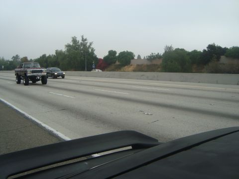 1986 Ford Bronco - A lifted brown pickup truck travels down the highway on a cloudy day, with other vehicles in the background.