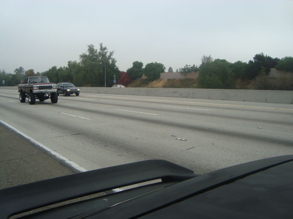 Brown lifted pickup truck driving on highway, front 3/4 view. Other vehicles and trees visible