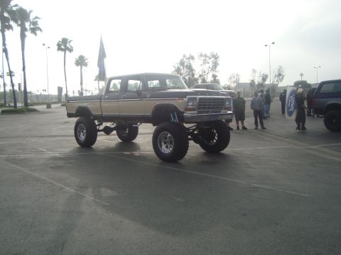 1986 Ford Bronco - A heavily modified pickup truck is parked in a lot with other vehicles and people nearby.