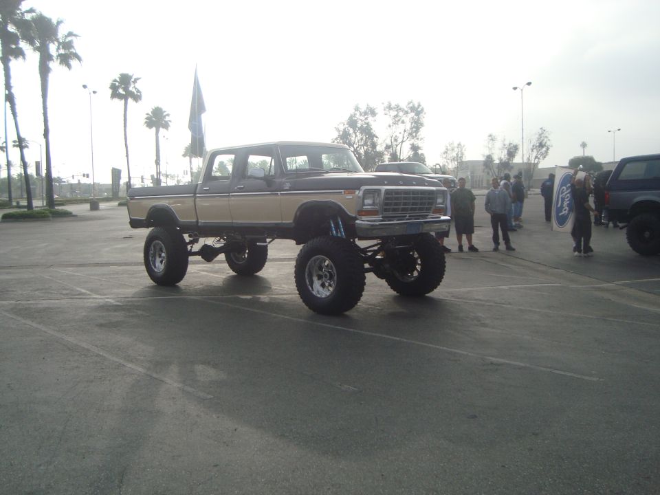 Lifted pickup truck with large tires, two-tone paint, and custom suspension. Other vehicles and people in background
