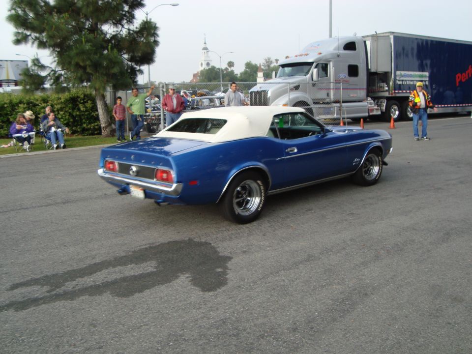 Blue convertible classic car parked on asphalt with a semi-truck behind it. People are sitting in chairs to the left