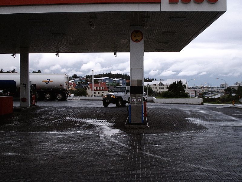 White soft-top 4x4 at Esso gas station pump with tanker truck and town in background