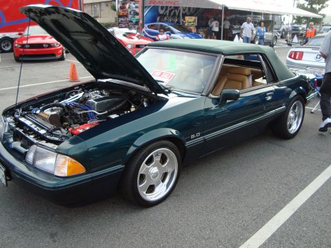 1986 Ford Bronco - A dark green convertible Mustang is parked with its hood up, revealing the engine bay. Other vehicles and tents are visible in the backgrou…