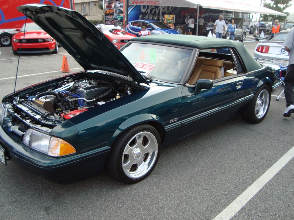Green convertible Mustang with hood open, showing engine. Other cars and tents in background