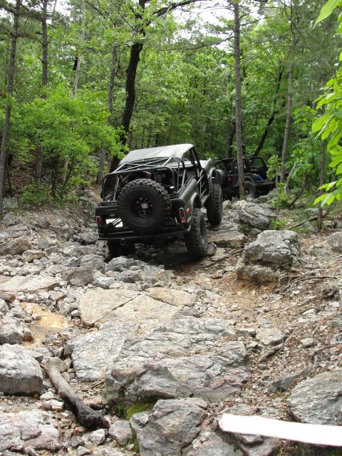1967 Ford Mustang - Two off-road vehicles tackle a challenging, rocky path through a dense forest.