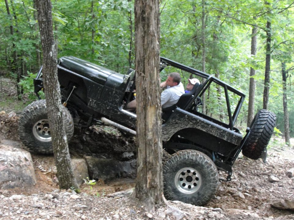 Black Jeep Wrangler with large tires navigating a steep, rocky incline in a wooded area. Two people are inside