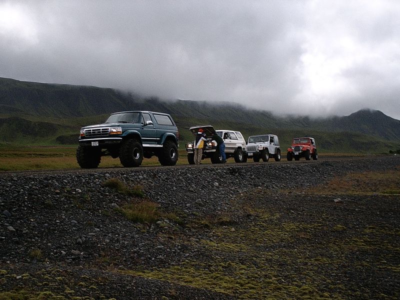 Lifted green Ford Bronco with large tires parked ahead of three 4x4s on a gravel road under misty mountains