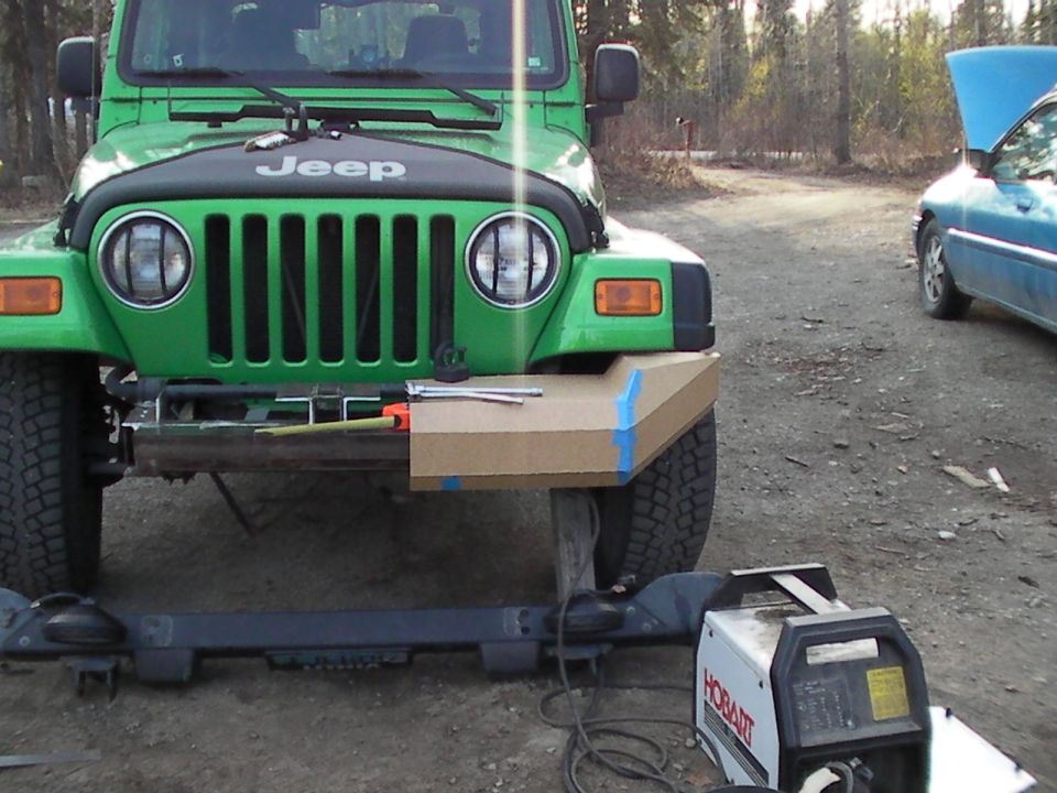 Green Jeep Wrangler front end with cardboard template for custom bumper, welding equipment nearby. Blue car in background