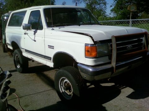 1989 Ford Bronco - Driver-side three-quarter front view of a lifted white Ford Bronco with a removable hardtop, fitted with a rusty tube grille guard, chrome…