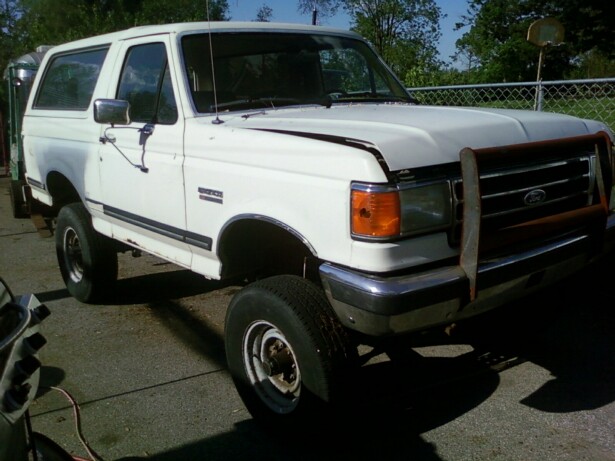 Lifted white Ford Bronco with tube grille guard, driver-side front view by a fence