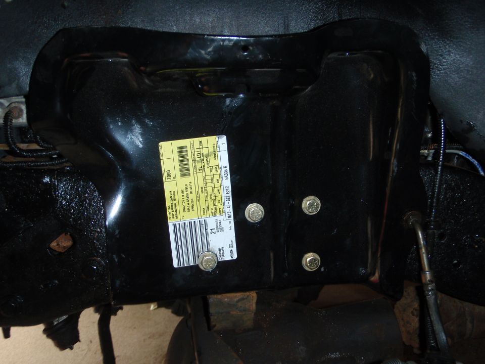 Black metal vehicle part with yellow sticker showing text and barcode. Several bolts are visible