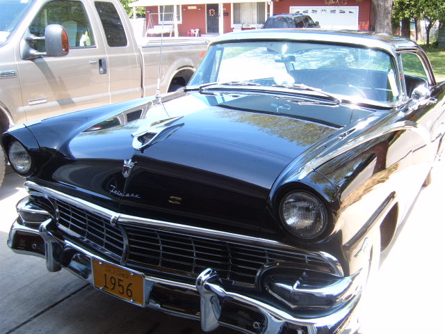 Black 1956 Ford Fairlane parked in front of a house with a pickup truck next to it