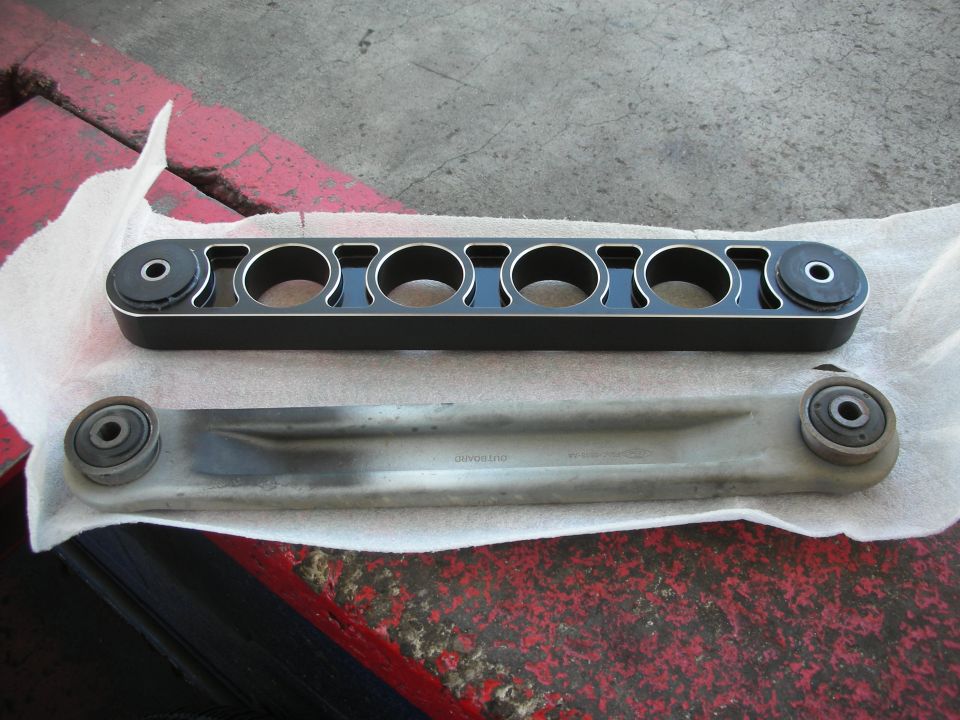 Aftermarket black control arm with six circular cutouts next to a used silver control arm on a white paper towel