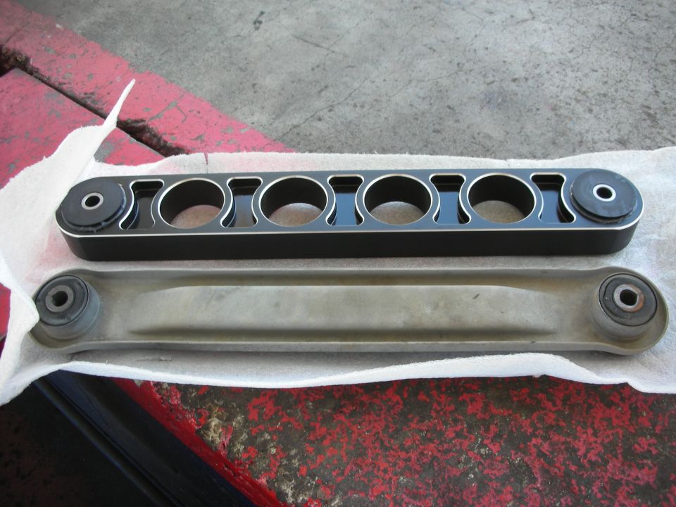Aftermarket black control arm with five circular cutouts next to a stock silver control arm