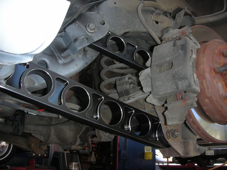 Aftermarket rear control arms with large circular cutouts installed on a vehicle's suspension system