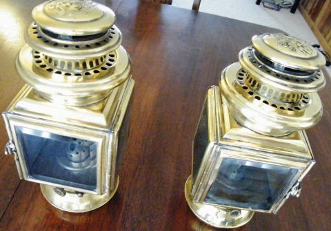 1912 Ford Model T - Two vintage brass carriage lamps are displayed on a wooden surface. The lamps feature intricate detailing on their tops and clear glass pan…