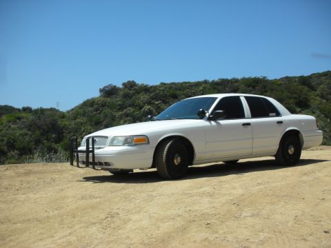 2004 Ford Crown Victoria - A white 2004 Ford Crown Victoria with a push bar is parked on a dirt road with a hilly, vegetated background.