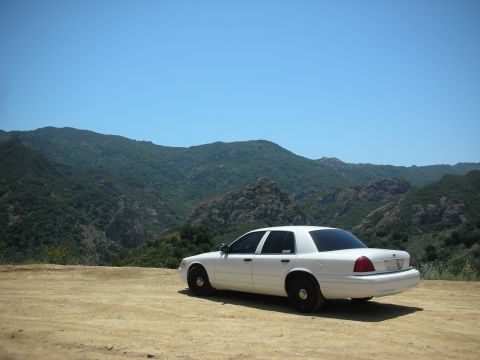 2004 Ford Crown Victoria - A white 2004 Ford Crown Victoria is parked on a dirt road with a mountainous landscape behind it.