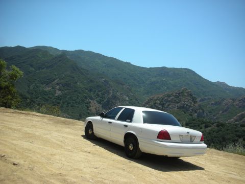 2004 Ford Crown Victoria - A white 2004 Ford Crown Victoria is parked on a dirt road, with a mountainous landscape behind it.