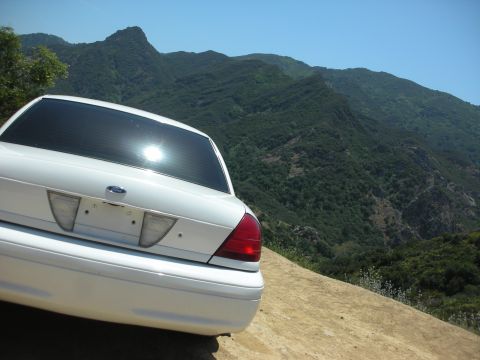 2004 Ford Crown Victoria - Rear view of a white 2004 Ford Crown Victoria parked on a dirt road with a scenic mountain backdrop.