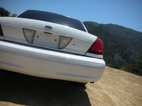 2004 Ford Crown Victoria - The rear of a white Ford Crown Victoria is parked on a dirt road with a mountainous landscape behind it.