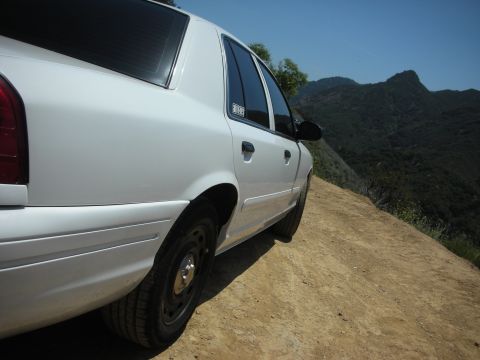 2004 Ford Crown Victoria - A white 2004 Ford Crown Victoria is parked on a dirt road overlooking a mountainous landscape.