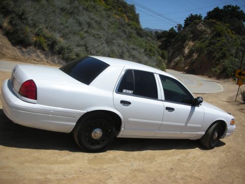 2004 Ford Crown Victoria - A white sedan is parked on a dirt shoulder beside a winding road in a hilly, vegetated area.