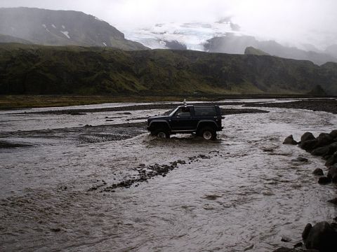 1996 Ford Bronco - A dark blue SUV navigates a wide, shallow river in a , mountainous landscape under an overcast sky.