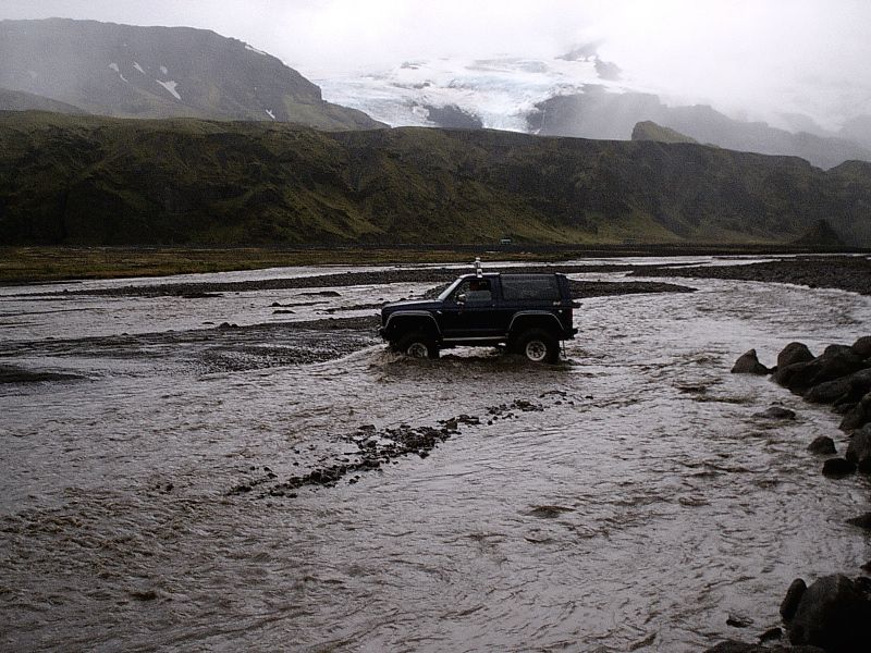Dark Ford Bronco SUV crossing a muddy glacial river below mountains and a glacier.