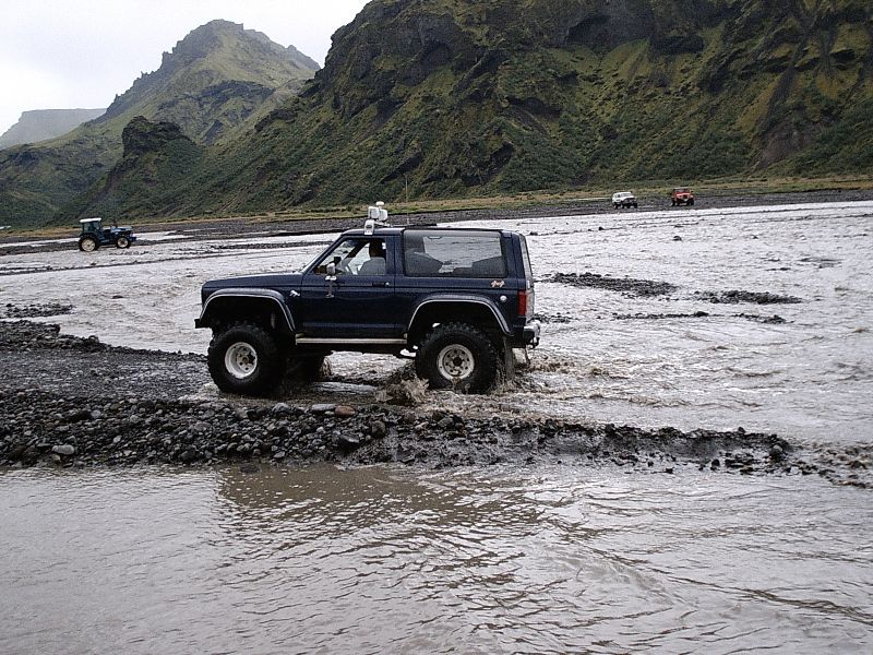 Dark blue lifted two-door Bronco crossing a wide glacial river with mountains behind.