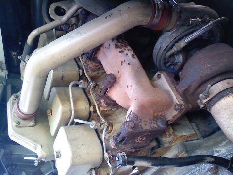 1990 Ford Bronco - Overhead close-up of the top of a tan-painted diesel engine showing a rusty cast-iron exhaust manifold feeding a turbocharger at right, wit…