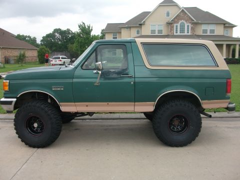 1989 Ford Bronco - Side view of a lifted green Ford Bronco with a tan removable top and large off-road tires.
