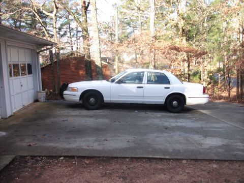 1999 Ford Crown Victoria - A white sedan is parked on a concrete driveway next to a garage with trees in the background.