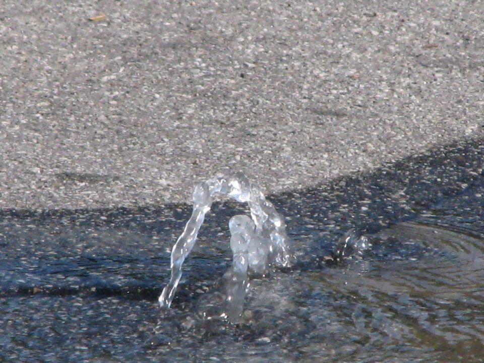 Water sprays from a hole in asphalt, creating a small fountain
