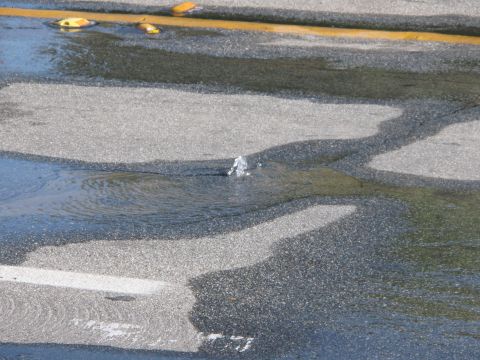1968 Ford Mustang - Water erupts from a broken water main on an asphalt surface, creating a puddle.