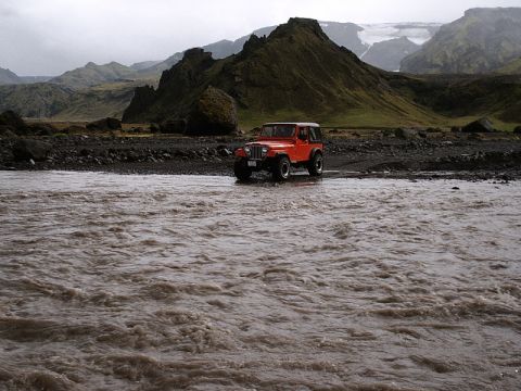 1996 Ford Bronco - An orange Jeep Wrangler navigates a turbulent, muddy river in a , mountainous landscape.