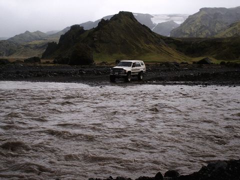 1996 Ford Bronco - A white SUV is parked on a rocky bank next to a turbulent river, with a dramatic mountain landscape behind it.