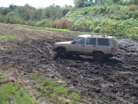 1992 Ford Bronco - A Jeep Cherokee XJ is stuck in thick mud on an off-road trail with vegetation in the background.