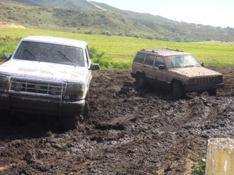 1992 Ford Bronco - Two SUVs are stuck in thick mud on a grassy hillside.