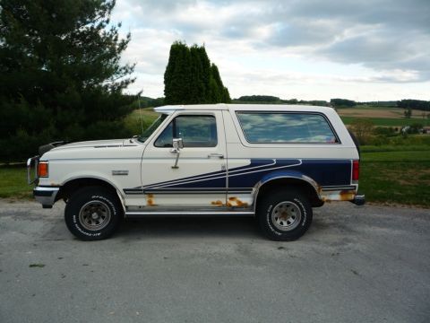 1988 Ford Bronco - White and blue Ford Bronco parked on asphalt with a rural landscape in the background.