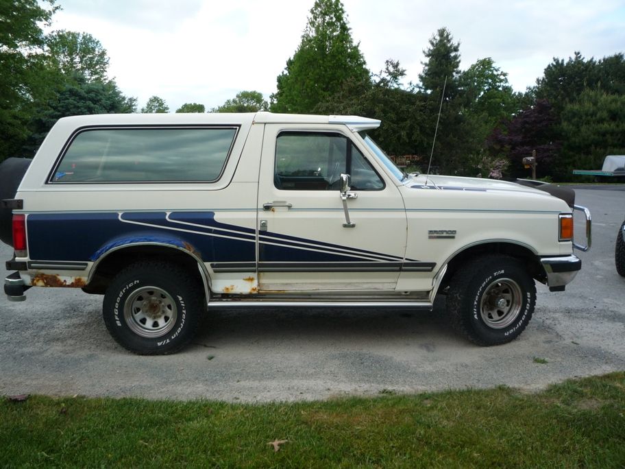 White and blue Ford Bronco with rust spots and a spare tire on the back, viewed from the side