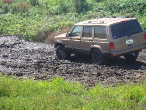 1992 Ford Bronco - A tan Jeep Cherokee XJ navigates a muddy terrain, its tires partially submerged.