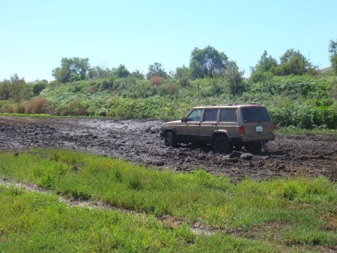 1992 Ford Bronco - Jeep Cherokee XJ off-roading through a muddy terrain under a clear blue sky.