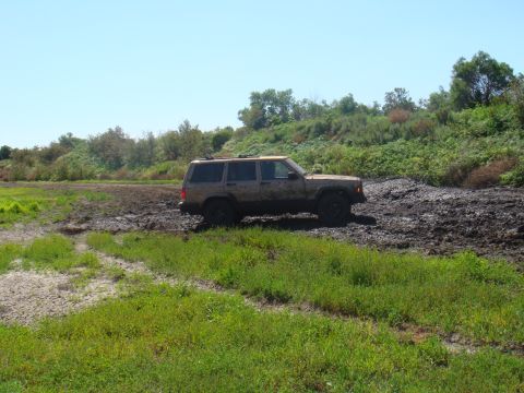 1992 Ford Bronco - A Jeep Cherokee XJ navigates a muddy off-road trail under a clear blue sky.