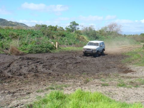 1992 Ford Bronco - A white SUV navigates a challenging muddy terrain on a dirt road under a bright blue sky.