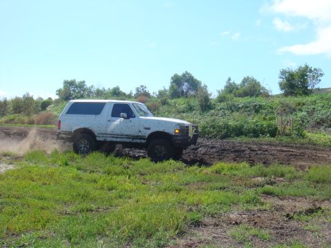 1992 Ford Bronco - A white 1992 Ford Bronco navigates a muddy off-road course under a clear blue sky.