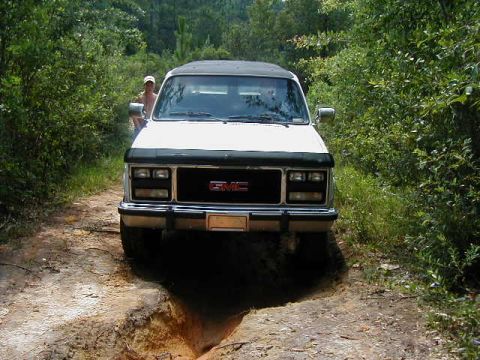 1990 Ford Bronco - A white GMC SUV navigates a muddy path surrounded by dense foliage.