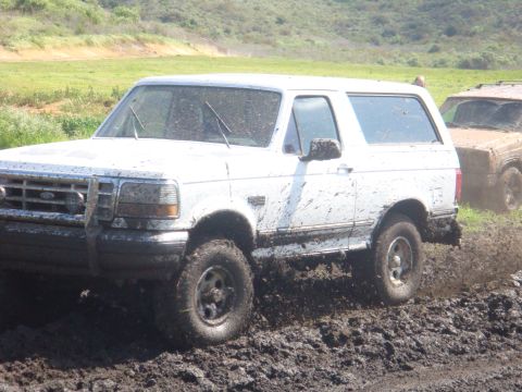 1992 Ford Bronco - A white 1992 Ford Bronco navigates a muddy terrain, with another vehicle partially visible in the background.